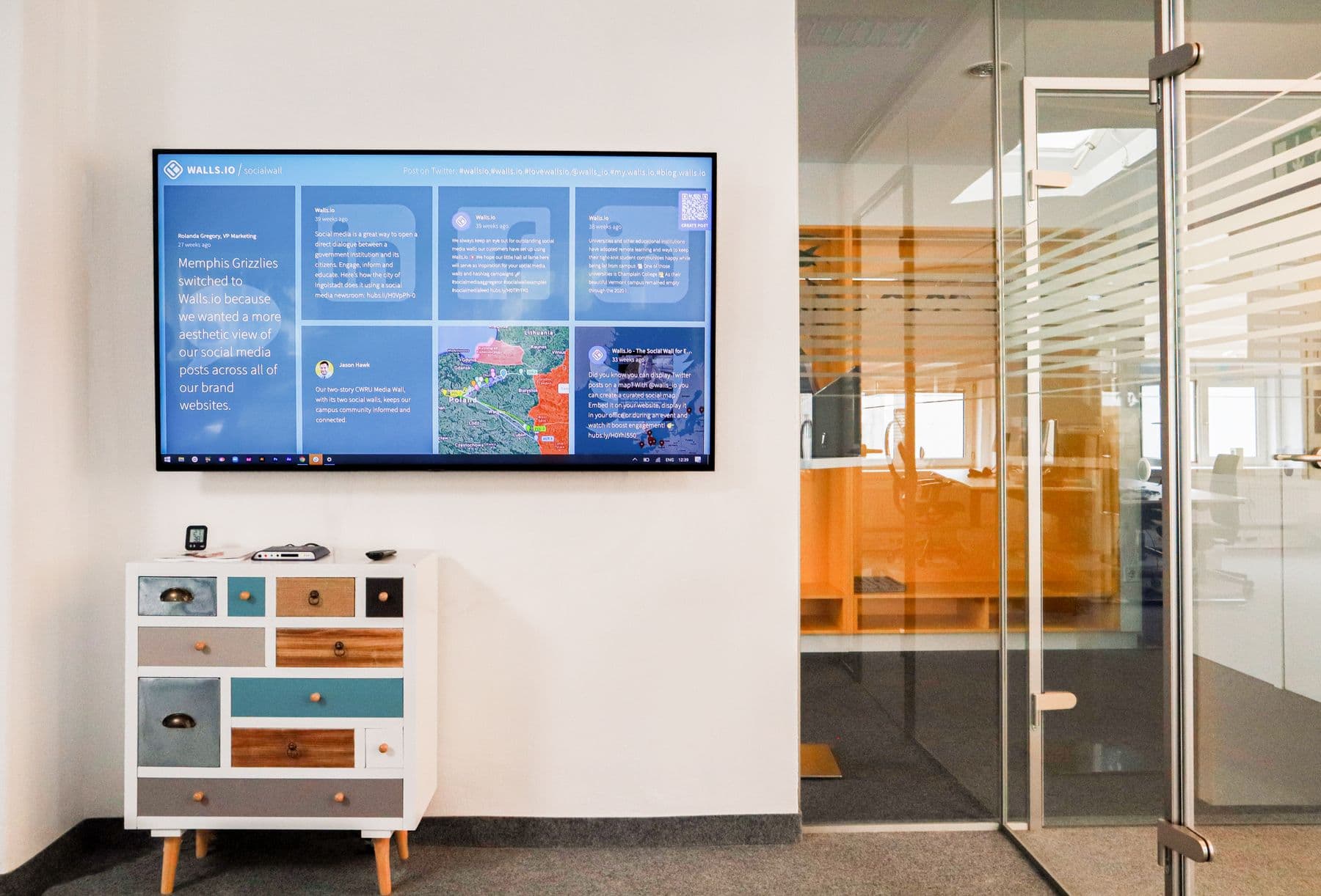 Office space with a wall-mounted display showing a social media dashboard beside glass partition walls