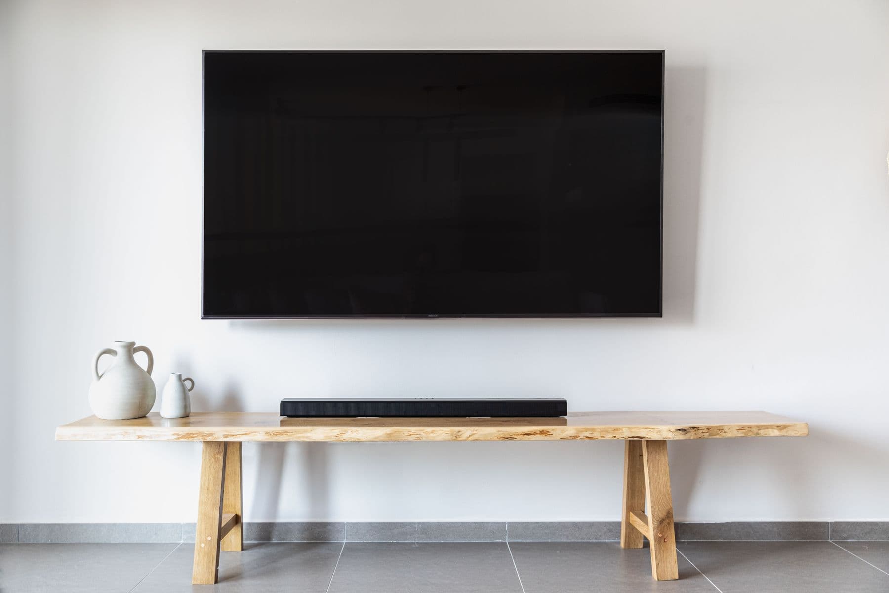 Large TV mounted flush on a white wall above a live-edge wood bench with a soundbar and ceramic vases